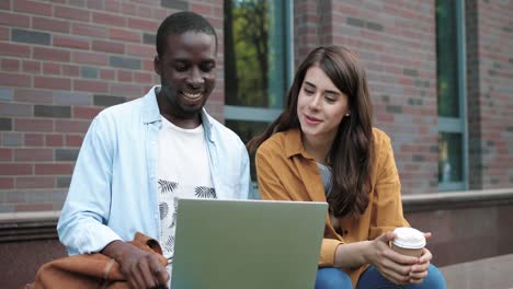 hombre afroamericano y mujer caucásica sentados en la calle y haciendo una videollamada en una laptop cerca de la universidad