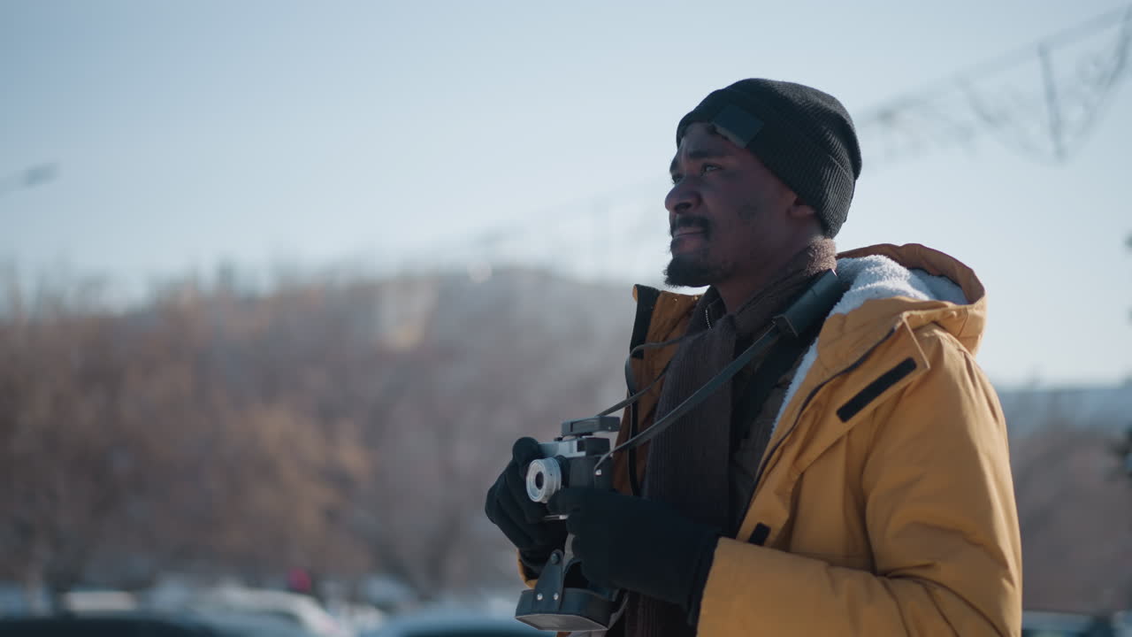 side view of black man in yellow coat and black beanie wearing gloves walking snowy urban street holding vintage camera and scanning surroundings under clear winter sky with parked cars