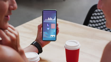 hombre y mujer caucásicos sentados a la mesa, usando un teléfono inteligente con estadísticas en la pantalla