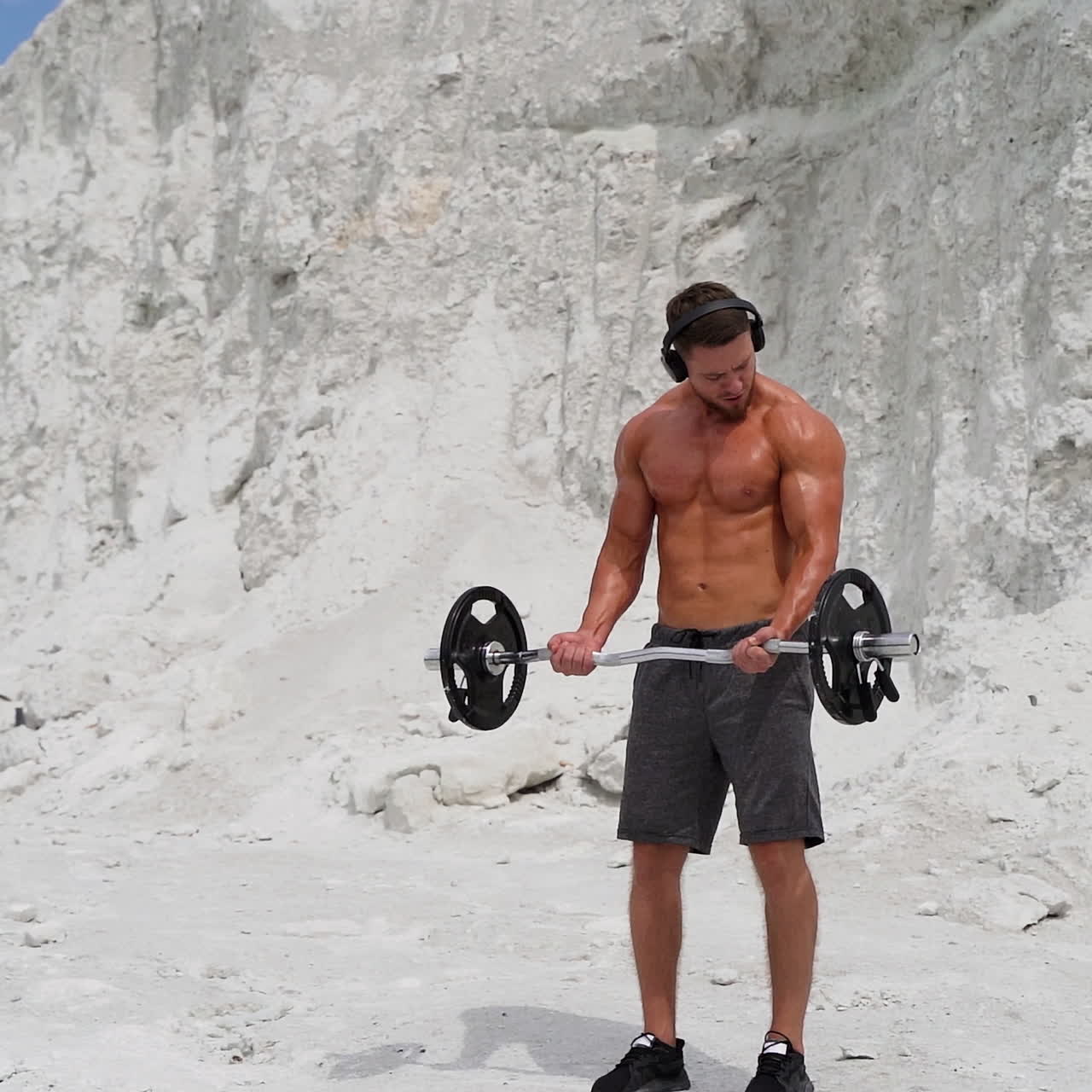 Muscular bodybuilder lift barbell on white mountain background. Strong young athlete with headphones doing his workout outdoors. Slow motion.
