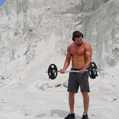 muscular bodybuilder lift barbell on white mountain background. strong young athlete with headphones doing his workout outdoors. slow motion.
