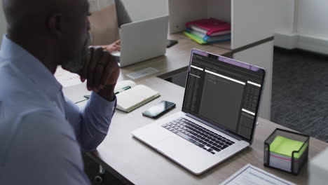hombre afroamericano sentado en el escritorio viendo el procesamiento de datos de codificación en la pantalla del portátil