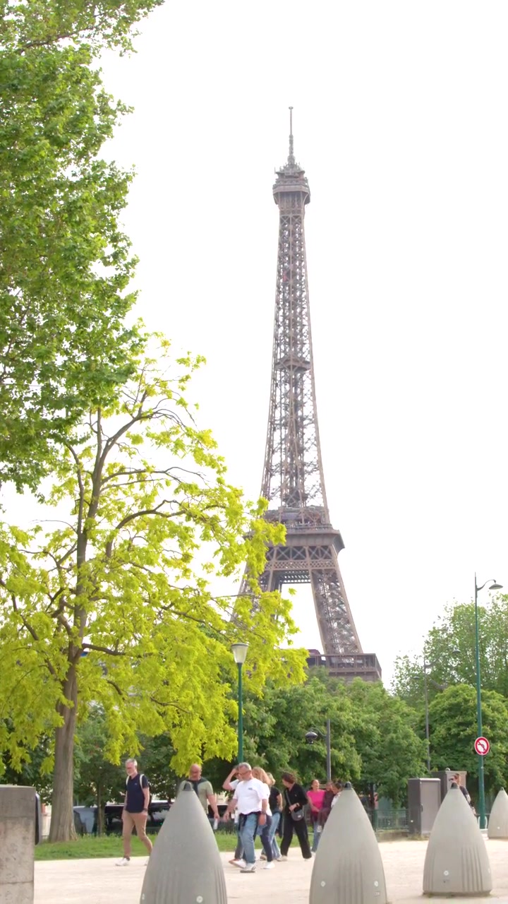 Inquadratura fissa e ampia di persone che passeggiano in un parco parigino con l'iconica Torre Eiffel.