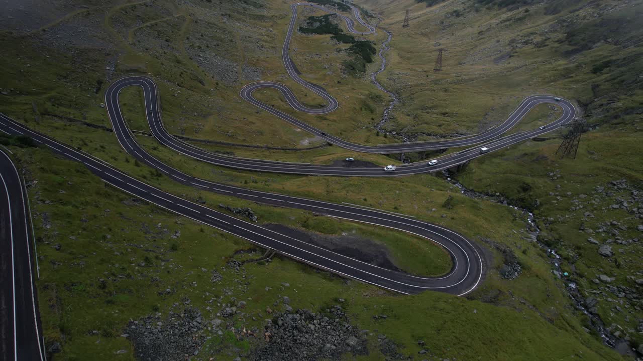 A forward-moving reveal shot of the Transfagarasan road, winding through a lush green valley, slowly unveiling the dramatic mountain scenery, with clouds drifting across the landscape.