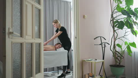 massage therapist wearing black outfit massaging client back in bright peaceful wellness room with gray curtain, indoor plant, open door and soft natural light contributing to calm relaxing ambiance