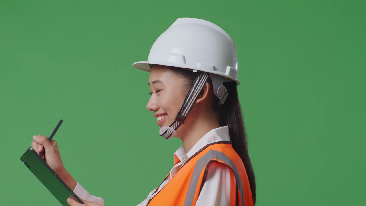 안전 헬을 입은 아시아 여성 엔지니어가 녹색 스크린 배경 스튜디오에서 걸어가는 동안 태블릿에 메모를 취하고 있습니다.