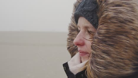 Young-woman-standing-in-the-wind-of-a-barren-landscape,-protected-from-the-freezing-cold-with-a-fur-hood