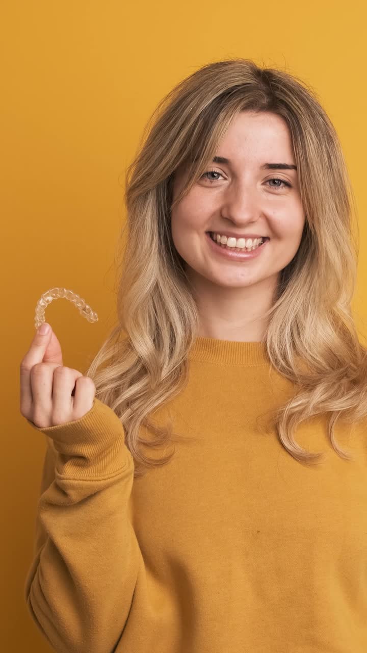 Cheerful young woman showing invisible tooth aligner in hand on yellow background