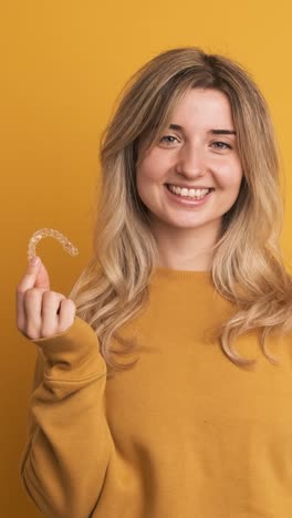 cheerful young woman showing invisible tooth aligner in hand on yellow background