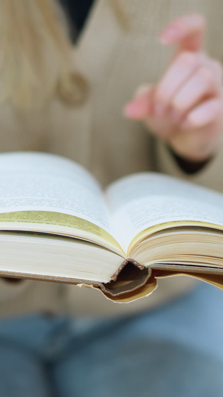 primer plano de una mujer con un suéter tejido de oro volviendo a la siguiente página del libro, mano en la página, flor decorativa verde ligeramente borrosa en el fondo