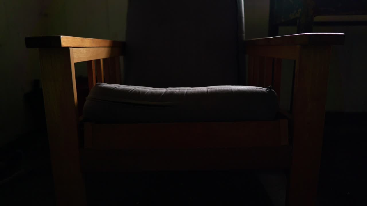 una toma de inclinación lenta revela un viejo sillón acolchado sentado en una habitación oscura e iluminado por rayos de luz de una ventana cercana