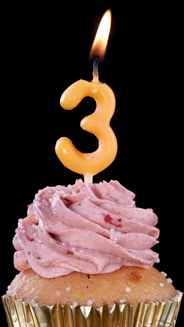 Una candela accesa con il numero tre brucia lentamente sopra un cupcake glassato alla fragola con pirottino dorato, su uno sfondo nero con illuminazione costante e senza movimento della telecamera