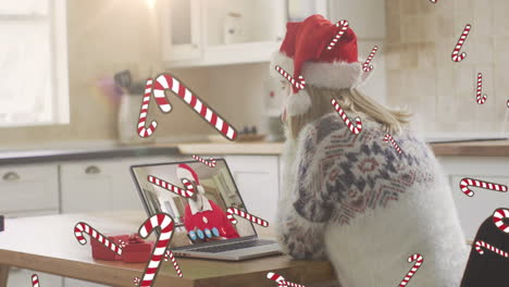 animación de bastones de caramelo cayendo sobre una mujer caucásica en sombrero de santa en una llamada de video portátil en navidad