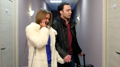 young man walking along hotel hall with suitcase while his girlfriend talking on the phone