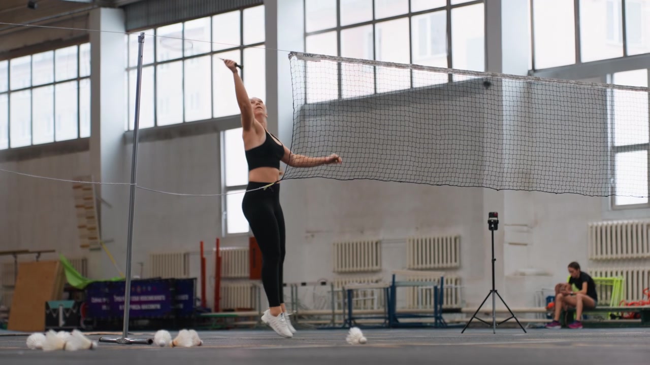 Donna sportiva che si prepara per uno scambio di volano. Donna atletica in abbigliamento nero che si riscalda con esercizi di volano. Atleta donna in una palestra vivace che dimostra tecniche precise di servizio e gioco di gambe.
