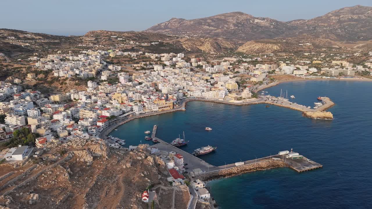 Drone performs a panoramic orbit over Pigadia port in Karpathos, revealing boats, coastline, and vibrant harbor life under clear blue skies