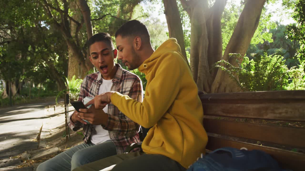 dos felices amigos de raza mixta sentados y usando un teléfono inteligente en el parque con mochilas