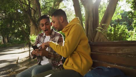 Two-happy-mixed-race-male-friends-sitting-and-using-smartphone-in-park-with-backpacks