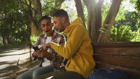 dos felices amigos de raza mixta sentados y usando un teléfono inteligente en el parque con mochilas
