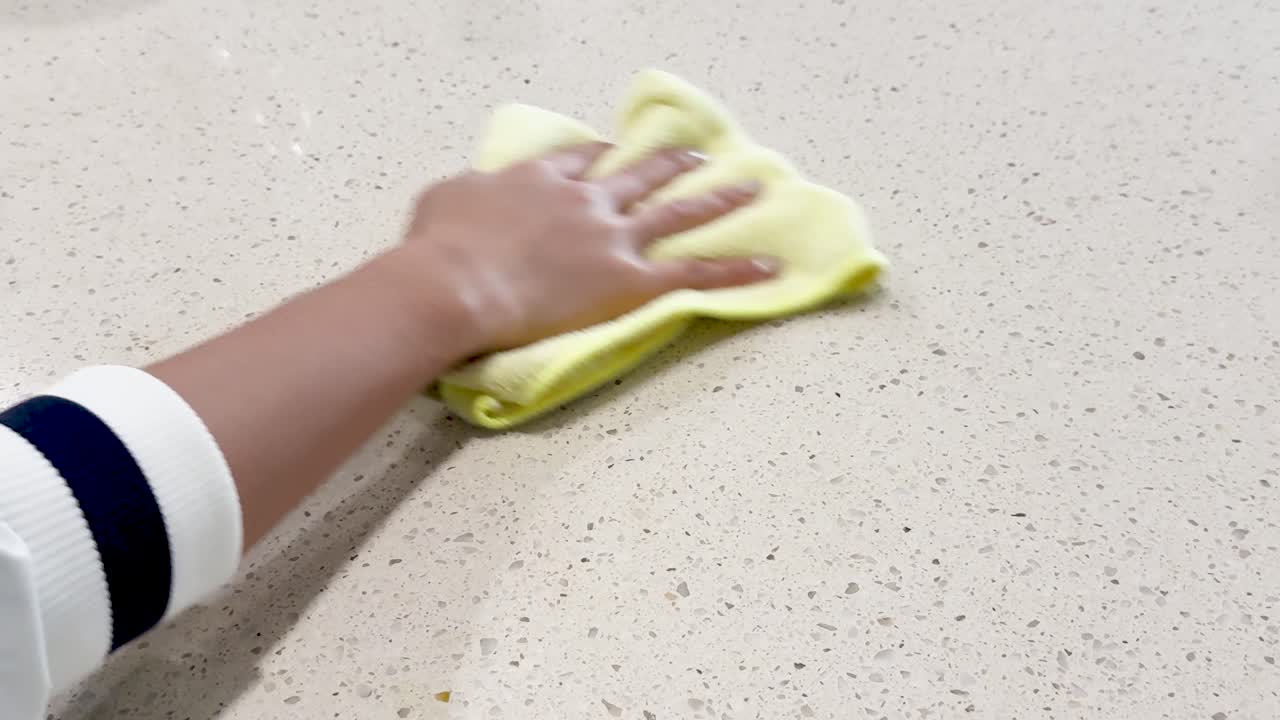 A hand wipes a speckled surface with a yellow cloth under bright lighting, emphasizing cleanliness and care