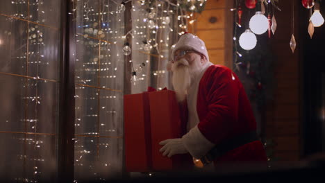 Santa-Claus-at-Christmas-stands-with-a-gift-in-his-hands-at-the-entrance-to-the-house-decorated-with-garlands-looking-out-the-window