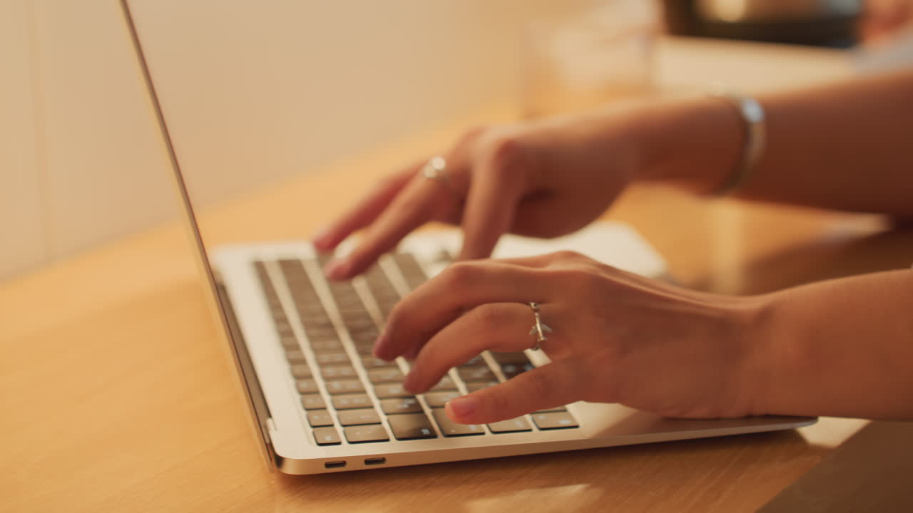 Close-up van handen die typen op een laptop, scène aan een hotelbureau met warm omgevingslicht van een lamp en een ondiepe scherptediepte (bokeh), een zichtbare trouwring om de vinger, geconcentreerd werken op afstand aan het opstellen van een e-mail en reisschema, een freelance schrijver.