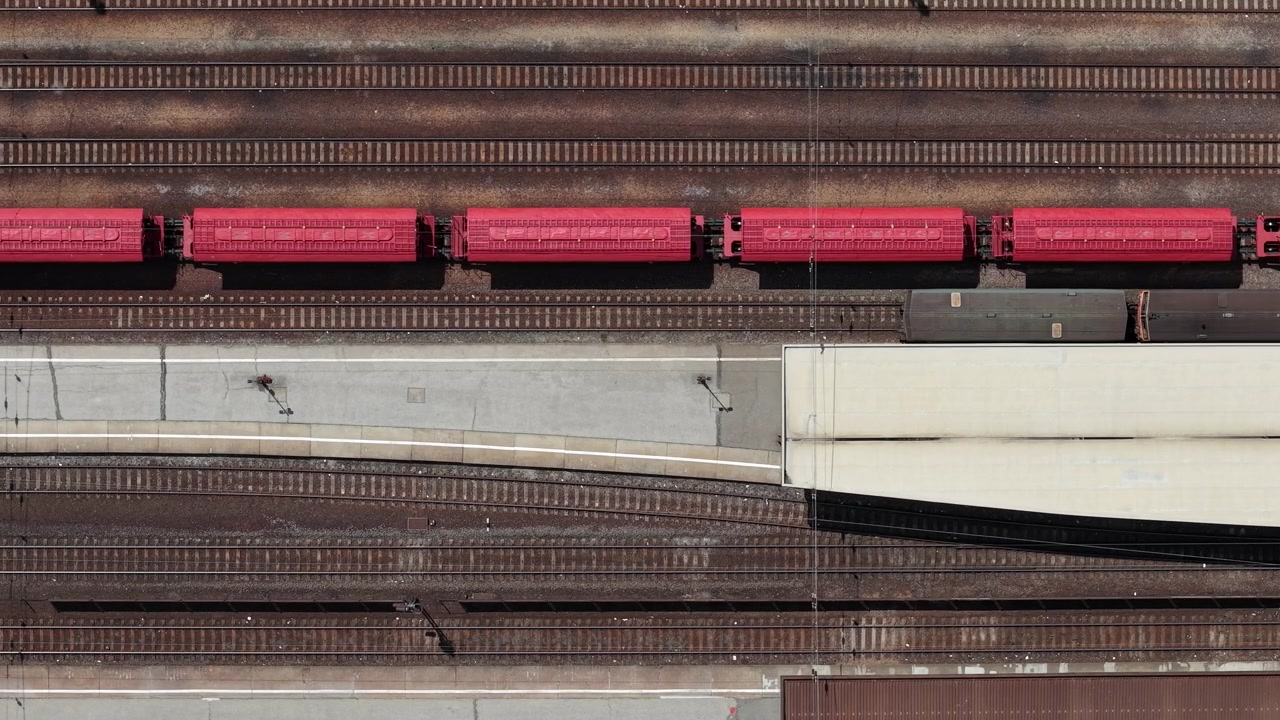 An aerial top-down view captures a vibrant red freight train traveling past a quiet railway platform in Hungary, industrial scenery.