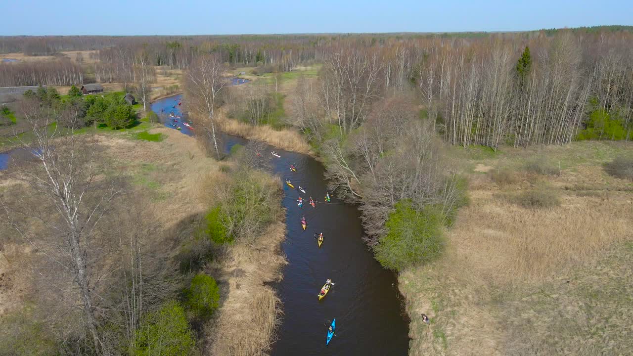 Aerial drone footage flying backwards over kayaks, paddleboards and boats that row on murky brown dark river water during summer time sunny day in Võhandu Marathon. Grassy shoreline and leafless trees