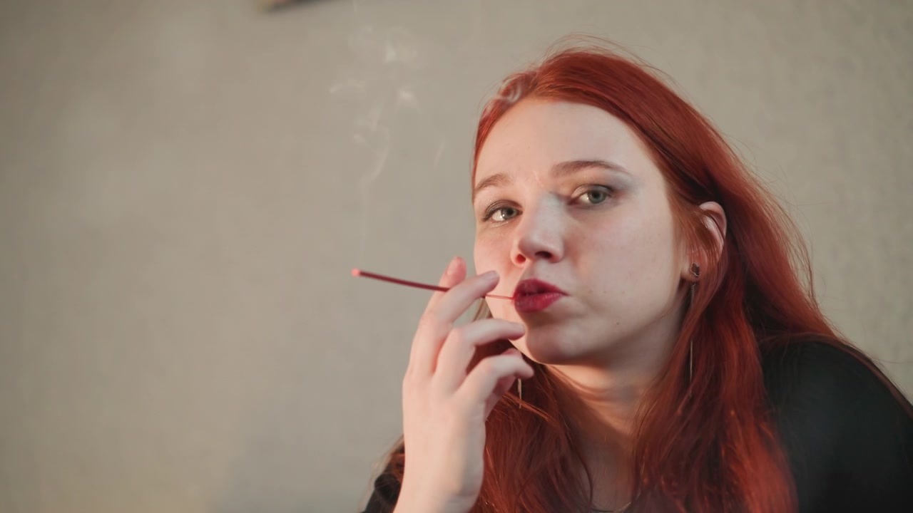 Junge Rothaarige raucht eine lange Zigarettenspitze, stimmungsvolles Innenporträt einer nachdenklichen Wahrsagerin, die Räucherstäbchenrauch bläst, Vintage-Make-up und Eyeliner, Tarot-Andeutungen außerhalb des Bildausschnitts, warme, weiche Beleuchtung.
