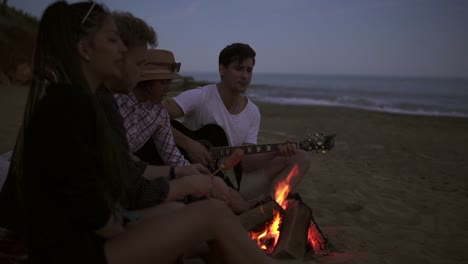 group of young and cheerful people sitting by the fire on the beach in the evening, grilling sausages and playing guitar