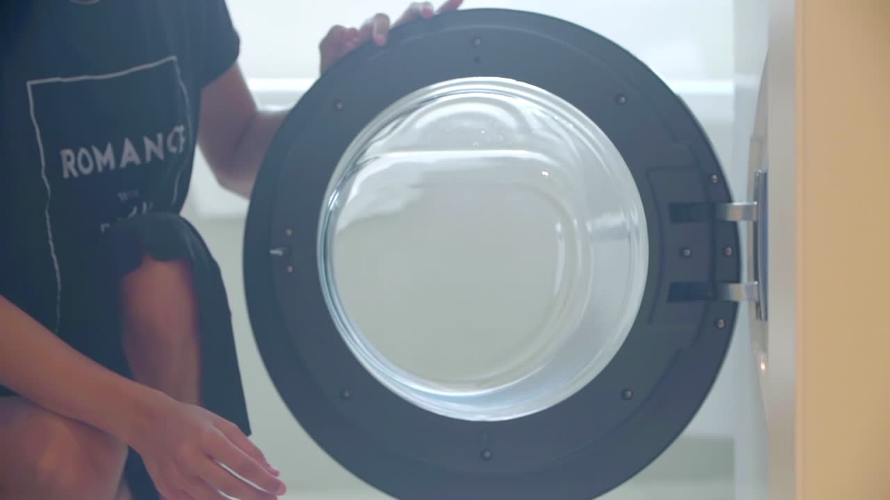 una mujer sexy en un vestido negro pone la ropa sucia en la lavadora y luego cierra la puerta