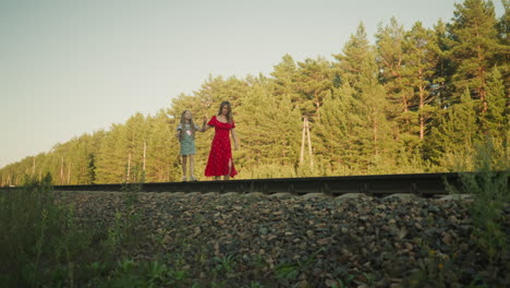 woman in red dress walking beside young girl on rail track surrounded by dense pine trees, holding hands as they move carefully together across rural area during peaceful golden hour moment
