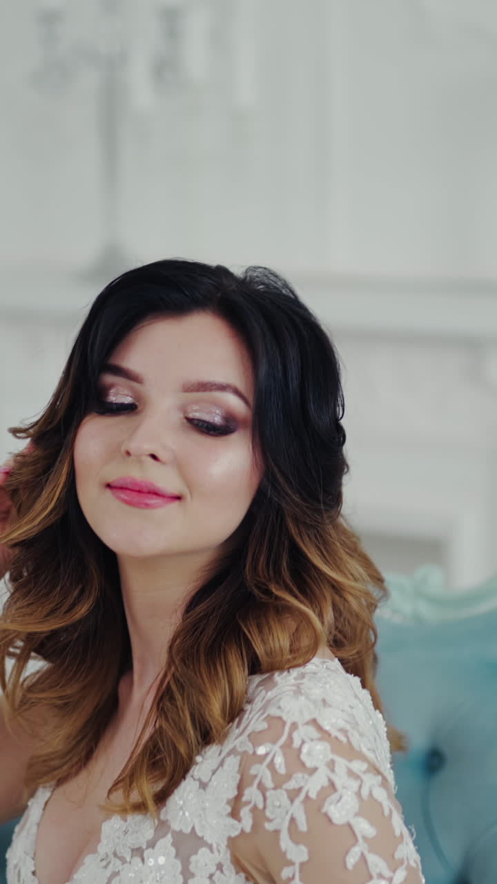 Incredible young woman in a beautiful white wedding dress is sitting on the couch. Lovely model with delicate makeup posing on royal sofa and smiling to camera. Vertical video