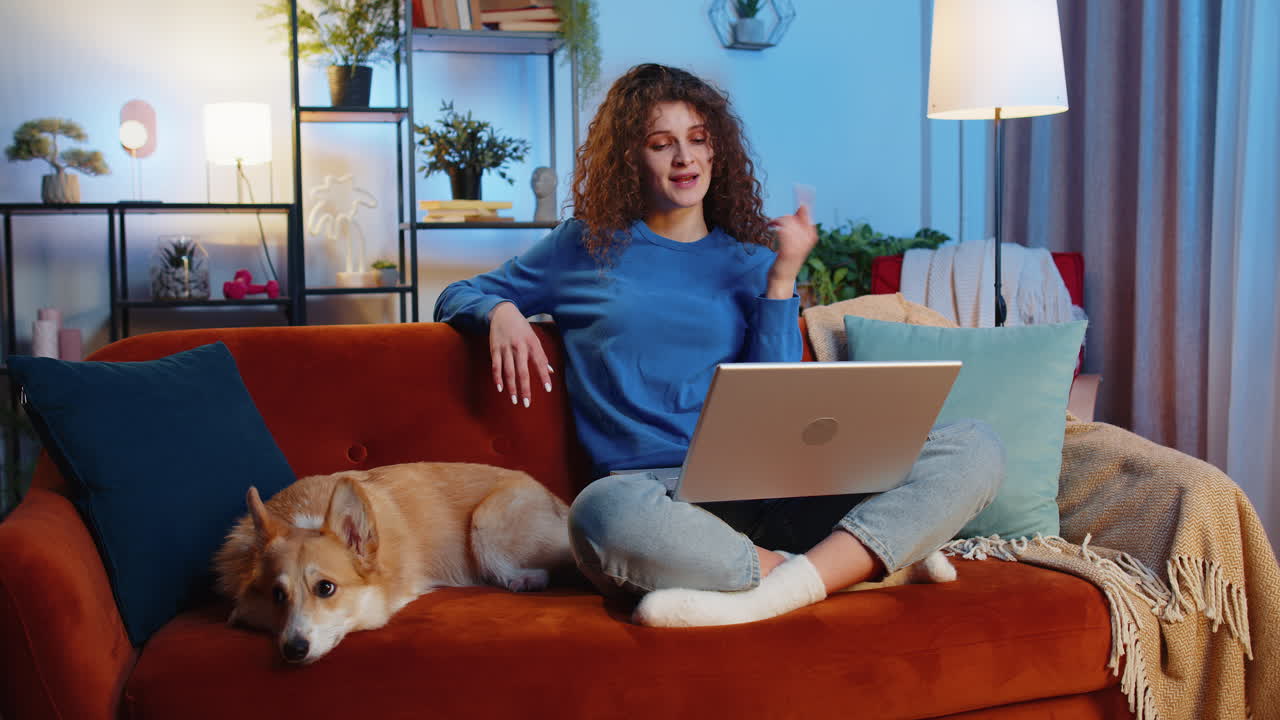 Smiling woman making video laptop webcam conference call while sitting beside corgi dog on home sofa