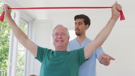 Senior-man-grimacing-while-exercising-with-resistance-band-against-male-therapist-at-nursing-home