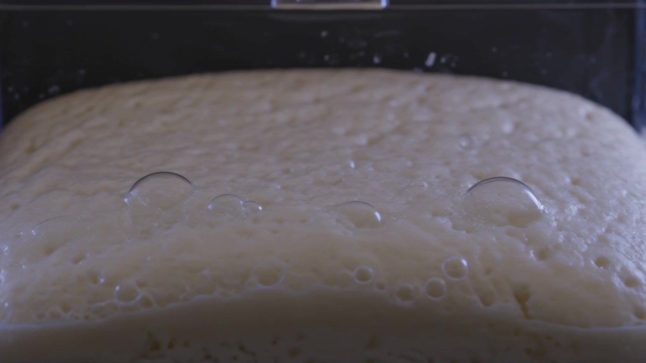 Mostrando impasto pallido in fermentazione che si espande in uno stampo da pane trasparente sul bancone della cucina, con il lievito che forma delle cupole