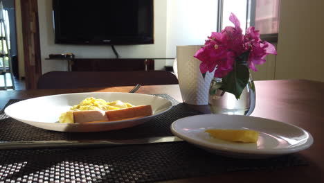 footage of breakfast sittings on the table with white plate white coffee mug flowers, scrambled eggs, fork and a knife with sunlight coming in from the window