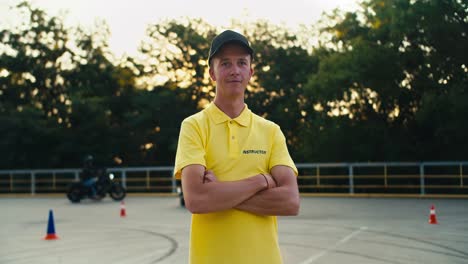 happy young driving instructor in yellow t-shirt. the guy poses with his arms folded on his chest against the background of green trees in a motorcycle school