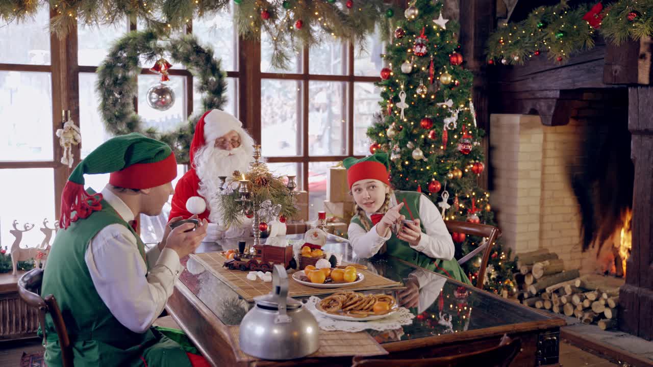 Santa Claus with elves. Cheerful elves sitting at the table together with Santa and drinking tea on Christmas background. Warm Christmas atmosphere.