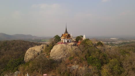 Aerial-footage-flying-backwards-revealing-a-Thai-Buddhist-temple-in-focus-also-showing-the-high-cliff-landscape-where-the-Wat-Phra-Phutthachai-temple-is-located,-Saraburi-,-Thailand