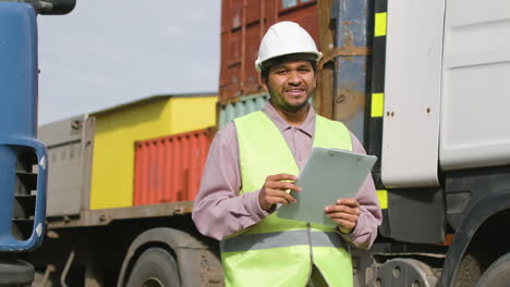 Arbeiter-Mit-Weste-Und-Schutzhelm,-Der-In-Einem-Logistikpark-In-Die-Kamera-Schaut-Und-Lächelt,-Während-Er-Dokumente-In-Der-Hand-Hält-1