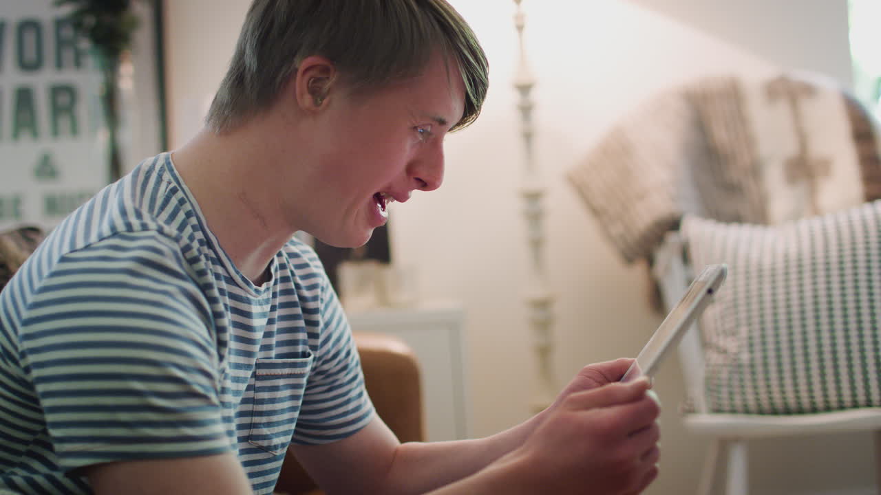 젊은 다운 증후군 남자가 소파에 앉아 집에서 디지털 태블릿을 보고 있습니다.