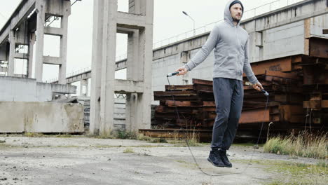 caucasian sportsman with beard in a grey hoodie jumping rope outdoors an abandoned factory on a cloudy morning