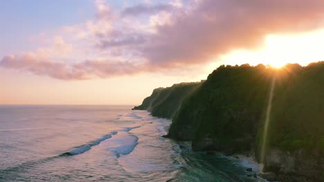 aerial of waves crashing onto uluwatu coastline with sun flare over cliffs at sunset in bali indonesia