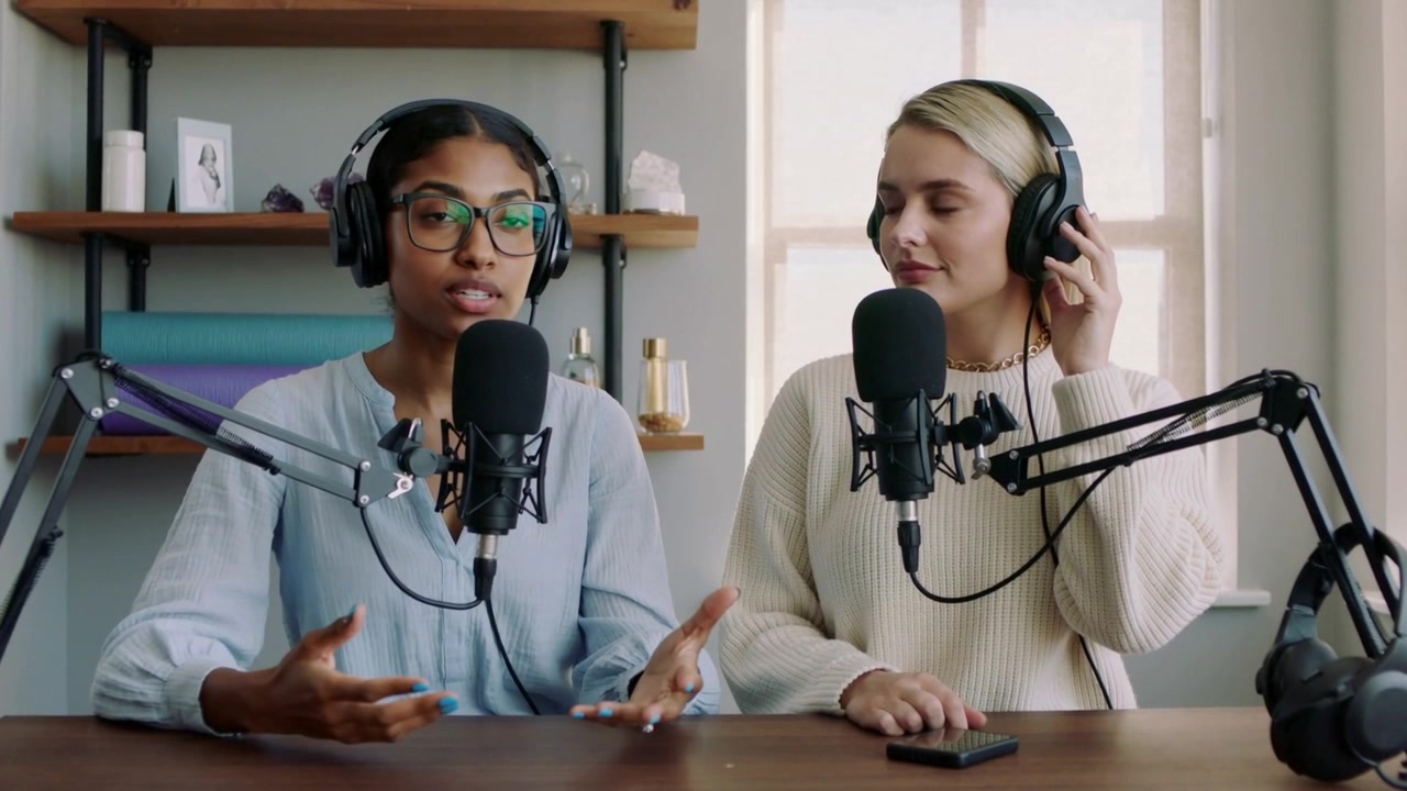 Speaking left host leading podcast in home studio, gesturing into mic, right host monitoring audio
