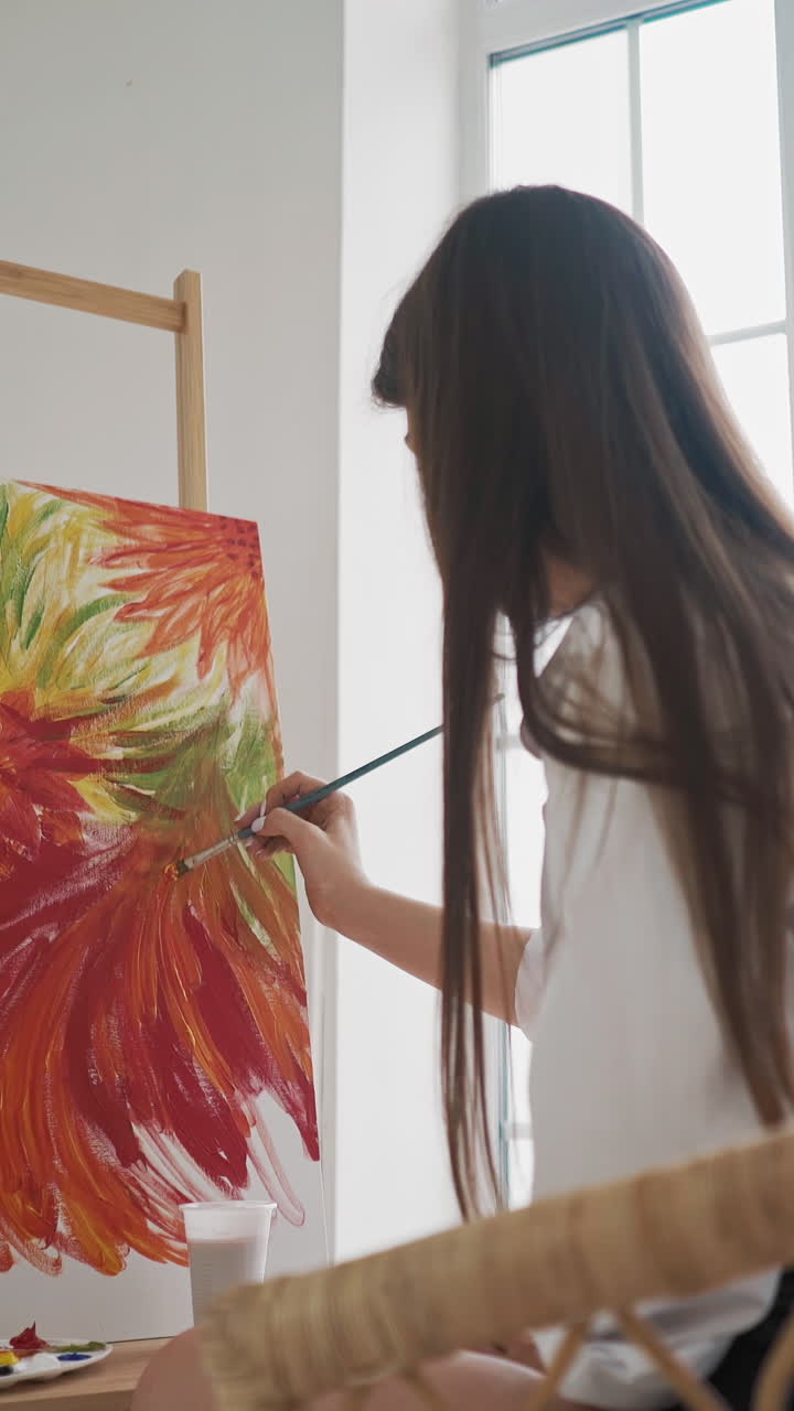 Long haired brunette lady artist at work on wonderful painting of large exotic flowers with red petals on canvas sits in armchair in studio backside view