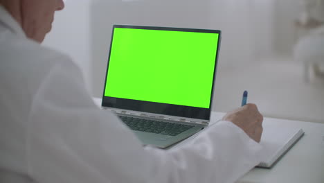 el médico está mirando la computadora portátil con pantalla verde en la mesa en la oficina de la clínica escribiendo notas