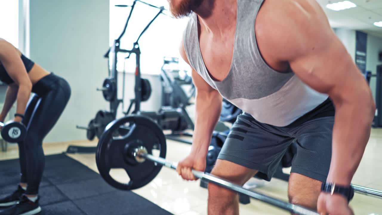 Athletic couple in sportswear lifting dumbbells in the gym. Persistent man is doing hard workout with dumbbells standing in special pose in the sports club.