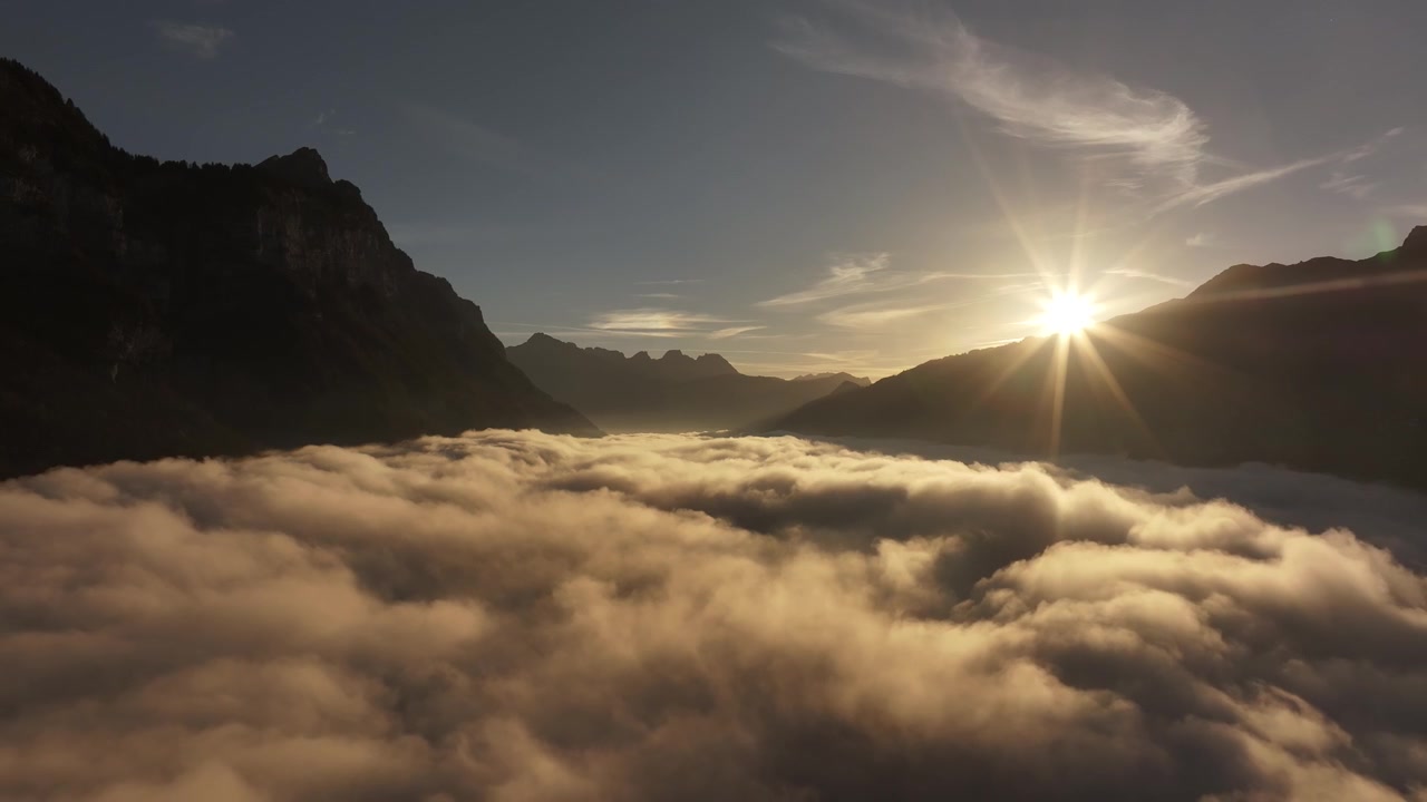 Drone che si allontana lentamente sopra uno strato di nuvole soffici annidato tra le Alpi svizzere, con la calda luce del sole mattutino che illumina delicatamente la scena
