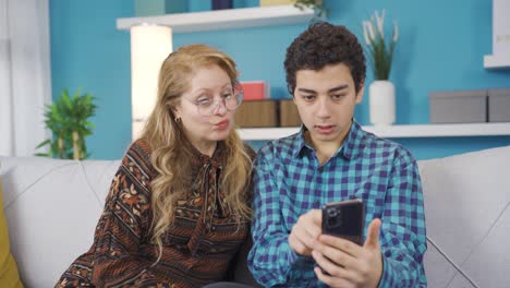 Un-Joven-Inteligente-En-Casa-Mostrando-A-Su-Madre-Cómo-Usar-Un-Teléfono-Móvil-Inteligente.